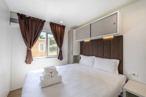A clean white bed with folded towels and pillows, brown curtains on a window, and overhead cabinets in a Desenzano Boutique Resor room