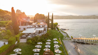 Desenzano Boutique Resort overlooking the lake with outdoor seating, white umbrellas, a pebble beach, and a wooden pier at sunset