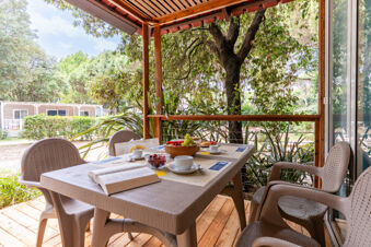 Outdoor covered wooden patio with table set for breakfast and surrounded by chairs and greenery at Jesolo Family Resort