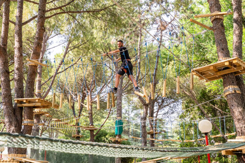 A man wearing black walks on a wooden rope bridge in a forest adventure park at Marina Julia Family Collection hotel