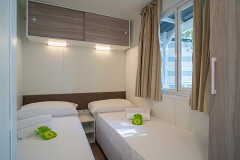 Two single beds with white linens and folded towels, a small nightstand, window with beige curtains in Marina Julia Family Collection room