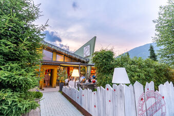 Outdoor dining area at Due Laghi Levico Family Collection with tables, chairs, people, white fence, and greenery surrounding the building