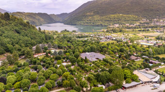 Aerial view of a green forested area with buildings, mountains, lake in background under cloudy sky at Due Laghi Levico Family Collection
