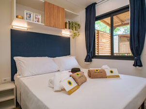 Bed with white sheets and pillows, folded towels with Club del Sole tags, wooden cabinets, and window with blue curtains at Due Laghi Levico Family Collection