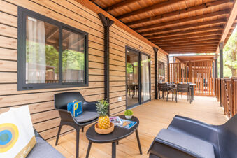 Outdoor wooden terrace with chairs and tables under a roof at Caldonazzo Family Collection with trees visible outside the windows