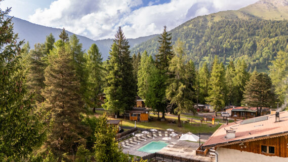 An outdoor pool with sun loungers and umbrellas surrounded by trees and wooden cabins at Val di Fiemme Easy Camping Village