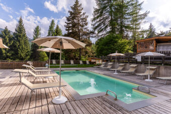 Outdoor swimming pool with beige lounge chairs and umbrellas surrounded by tall trees at Val di Fiemme Easy Camping Village