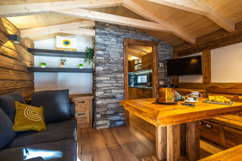Living area with dark sofa, wooden table and benches, stone wall, kitchen entrance, and TV at Val di Fiemme Easy Camping Village
