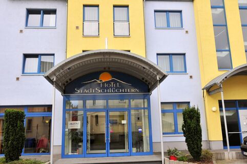 Entrance of Akzent Hotel Stadt Schl&uuml;chtern with blue framed glass doors, yellow and white facade, and two small trees outside