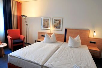 Two single beds with white linens and pillows, wooden headboards, a red armchair, floor lamp, and two paintings in Akzent Hotel Stadt Schl&uuml;chtern