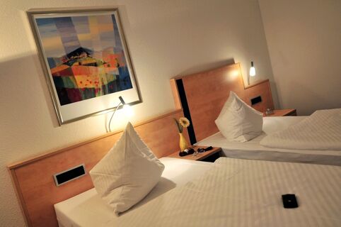 Two single beds with white linens, triangular pillows, wooden headboards, bedside lamps, and an abstract painting in Akzent Hotel Stadt Schl&uuml;chtern