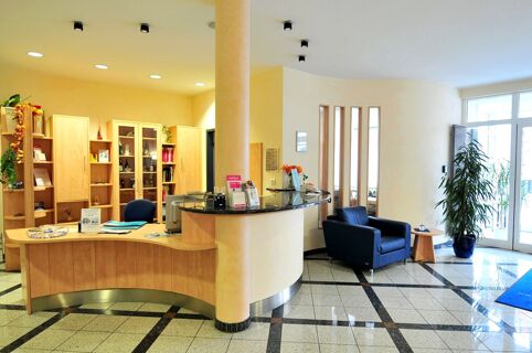 Reception area with a curved wooden desk, blue chair, bookshelves, a black armchair, small table, and plant at Akzent Hotel Stadt Schl&uuml;chtern