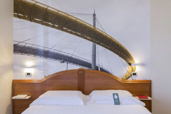Double bed with white linens and wooden headboard against a wall featuring a large bridge mural in B&B Hotel Pescara