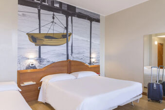 Hotel room at B&B Hotel Pescara with double bed, single bed, large wall photo of a yellow boat, suitcase, and full-length mirror