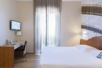 Minimalist hotel room at B&B Hotel Pescara with a made bed, a wooden desk with lamp and chair, and a window with sheer curtains