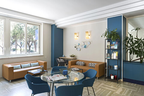 A modern seating area at B&B Hotel Pescara with leather sofas, blue armchairs, a glass table, decorative plants, and natural light from large windows