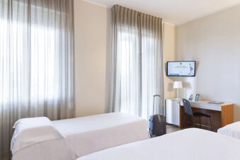 Hotel room at B&B Hotel Pescara with two single beds, gray curtains, suitcase, desk with chair, lamp, and wall-mounted TV
