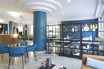 Bright and modern common area at B&B Hotel Pescara with blue chairs, round glass table, shelving unit, and a beige sofa with pillow