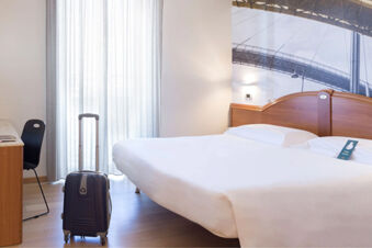 Hotel room at B&B Hotel Pescara with a double bed, a black suitcase, a chair, and a desk near a sheer curtained window