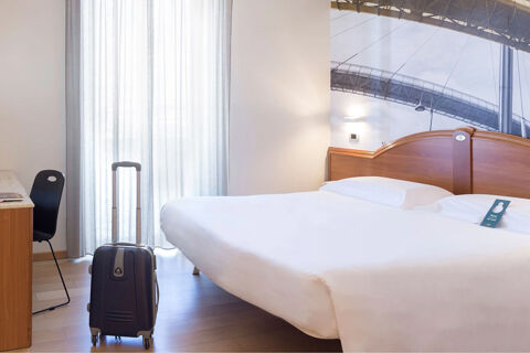 Hotel room at B&B Hotel Pescara with a double bed, a black suitcase, a chair, and a desk near a sheer curtained window