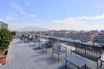 Outdoor terrace seating with black metal chairs, tables, railings, and potted plants overlooking the city at B&B Hotel Napoli