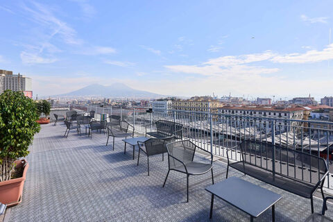 Outdoor terrace seating with black metal chairs, tables, railings, and potted plants overlooking the city at B&B Hotel Napoli