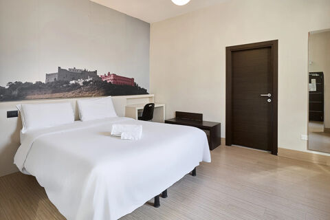 A double bed with white linens and two pillows in a room with a dark wood door, desk, chair, and mural at B&B Hotel Napoli