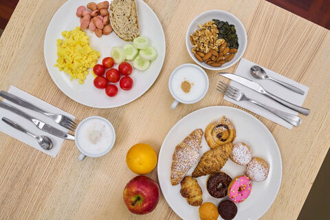 Two plates with breakfast foods including scrambled eggs, sausages, bread, vegetables, pastries, nuts, an apple, orange, and two cappuccinos at B&B Hotel Napoli