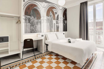Hotel room at B&B Hotel Napoli with white bedding, a mural of architectural arches, a desk with chair, and a window with gray curtains