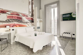 Bright hotel room at B&B Hotel Napoli with a white double bed, wall art, desk, chair, open closet, and TV mounted on wall