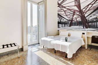 Bright hotel room at B&B Hotel Napoli with twin beds, white linens, a large window, decorative wall art, and a luggage rack