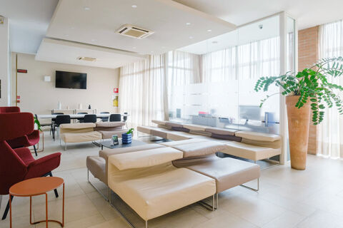 Bright waiting area at B&B Hotel Reggio Emilia Remilia with beige modern sofas, red chairs, large windows, and a tall plant