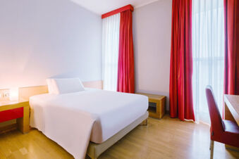 A minimalist hotel room at B&B Hotel Reggio Emilia Remilia with a white bed, light wood furniture, red curtains, and a red chair