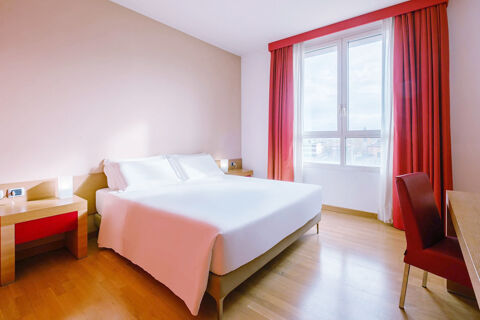 Minimalist hotel room at B&B Hotel Reggio Emilia Remilia featuring a large bed, desk with red chair, and window with red curtains