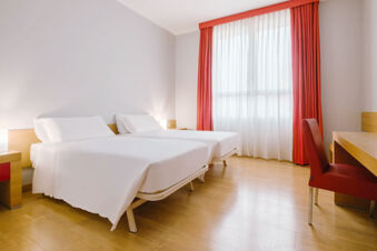 Two neatly made single beds with white linens in a bright room with wooden flooring and red curtains at B&B Hotel Reggio Emilia Remilia
