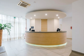 Modern circular wooden reception desk with computers beneath white ceiling lights in B&B Hotel Reggio Emilia Remilia lobby