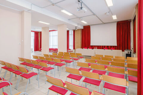 Conference room with rows of wooden chairs with red cushions, a projector, and a screen at B&B Hotel Reggio Emilia Remilia