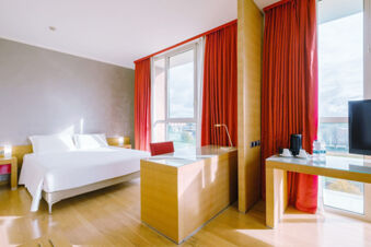 A bright hotel room at B&B Hotel Reggio Emilia Remilia with large windows, red curtains, a double bed, and modern wooden furniture