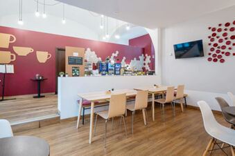 Dining area at B&B Hotel Trieste with wooden tables, chairs, a TV mounted on the wall, and a coffee station against a red wall