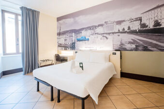 Bright hotel room at B&B Hotel Trieste with a double bed, white linens, large black and white wall photo, and window with curtains