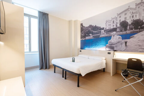 A neat hotel room at B&B Hotel Trieste with a double bed, a large window with gray curtains, and a desk with a chair