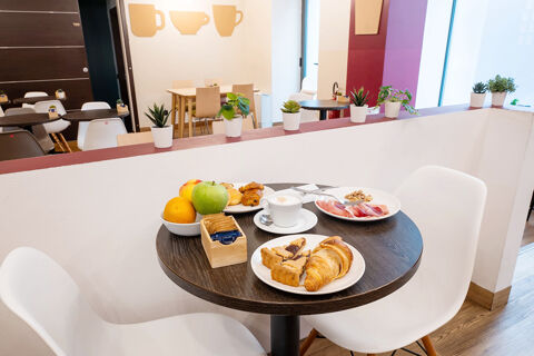 A small round table with two white chairs at B&B Hotel Trieste set with croissants, fruit, coffee, and plates of snacks in a cozy dining area