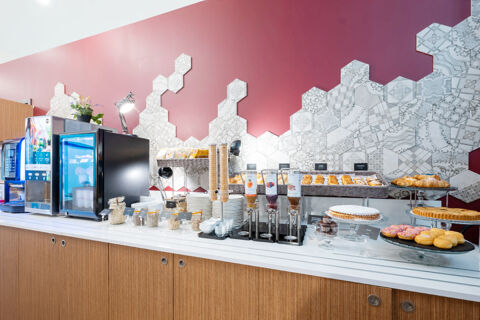 Breakfast buffet setup with pastries, juice dispensers, cereal jars, plates, and coffee machine at B&B Hotel Trieste