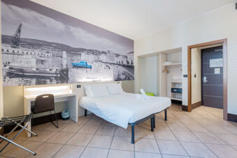 Hotel room at B&B Hotel Trieste with a double bed, desk and chair, open wardrobe, and a large black and white wall mural of the harbor