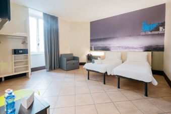 Hotel room at B&B Hotel Trieste with two single beds, a chair, desk, open wardrobe with safe, and a large window with grey curtains