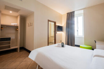 Hotel room at B&B Hotel Trieste featuring a bed with white linens, a wall-mounted TV, a closet with hangers and a safe, and a window with curtains