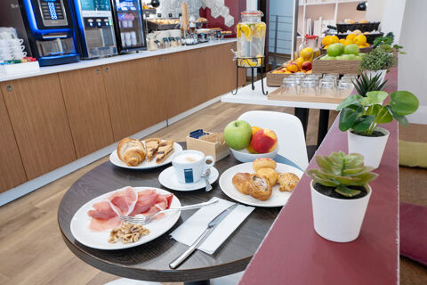 Table at B&B Hotel Trieste with plates of pastries, cured meat, fruit, a cappuccino, and utensil beside a breakfast buffet area