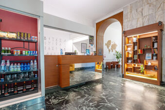 Reception desk and illuminated wooden shelving unit with books and plants in the lobby of B&B Hotel Trieste with a refrigerated display on the left