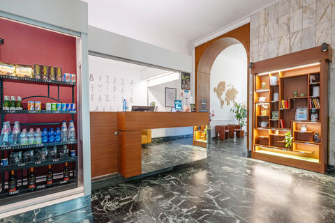 Reception desk and illuminated wooden shelving unit with books and plants in the lobby of B&B Hotel Trieste with a refrigerated display on the left