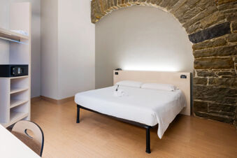 Minimalist hotel room at B&B Hotel Trieste with white bed linens, stone arch wall, wooden floor, and built-in shelving with a safe